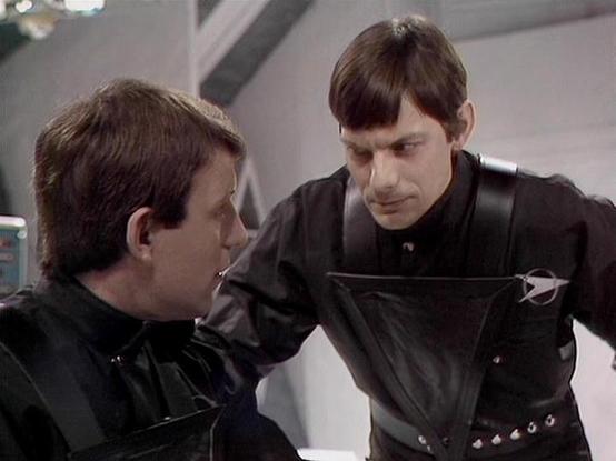 Claude Sonnet 4.5 describes the image as: "This image shows two actors in dark, military-style uniforms engaged in what appears to be an intense conversation. The setting has a futuristic, sterile appearance with grey walls and minimal decoration in the background, suggesting a science fiction production environment, likely a spaceship or advanced facility.

The person on the left wears a black uniform and looks toward their companion with concern or attention. The person on the right, dressed …