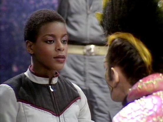 Claude Sonnet 4 describes the image as: "I can see this appears to be from a science fiction production, likely from the 1980s based on the visual style and futuristic costume design. The scene shows two people in conversation - one wearing a distinctive futuristic uniform with geometric patterns in dark and light colors with trim details. The setting appears to be indoors with atmospheric lighting. The production values and aesthetic suggest this is from a British science fiction series from t…