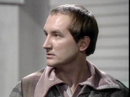 Claude Sonnet 4.5 describes the image as: "This image shows a profile shot of a male actor in what appears to be a scene from a 1970s or early 1980s television production. The actor is wearing a utilitarian, earth-toned costume consisting of a rust or taupe-colored jacket with a collar, suggesting a science fiction or military setting. The lighting and video quality are characteristic of television productions from that era.

The actor has dark hair styled in a period-appropriate cut and is gaz…