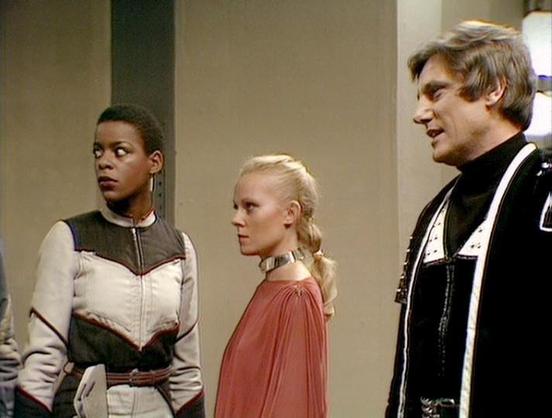 Claude Sonnet 3.7 describes the image as: "This image appears to be a scene from the science fiction series "Blake's 7," showing three characters in distinctive costume design typical of the show's futuristic aesthetic. 

The scene features three people standing together in what looks like a spacecraft or space station interior with neutral-colored walls. On the left is a person wearing a white and gray outfit with geometric patterns and a metallic collar. In the center is someone with blonde h…
