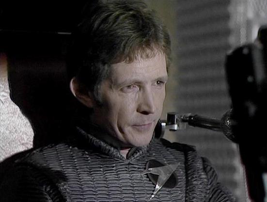 Claude Haiku 4.5 describes the image as: "# Image Description

This image appears to be a scene from a film or television production, shot in black and white or with muted color grading. The setting looks like an interior space, possibly a spacecraft or industrial facility, given the metallic and geometric architectural elements visible in the background.

The main subject is a man shown in profile, wearing a dark textured jacket or tunic with a quilted pattern. He appears to be in a tense or s…