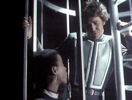 Claude Sonnet 4 describes the image as: "This image appears to be from a science fiction television production, likely from the 1980s based on the visual style and production quality. The scene shows two characters in what appears to be a spacecraft or futuristic facility, with metallic structural elements and technological equipment visible in the background. The lighting creates a dramatic atmosphere typical of sci-fi productions of that era.

One character is positioned lower in the frame, w…