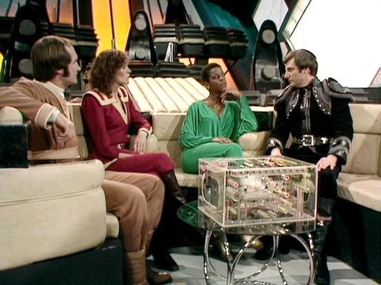 Claude Sonnet 4 describes the image as: "This appears to be a scene from the British science fiction television series "Blake's 7," likely from the late 1970s or early 1980s. The setting is the interior of what looks like a spaceship or futuristic facility, with characteristic sci-fi design elements including angular architecture and metallic surfaces visible in the background.

Four actors are seated in a comfortable lounge area with cream-colored furniture arranged around a distinctive glass …