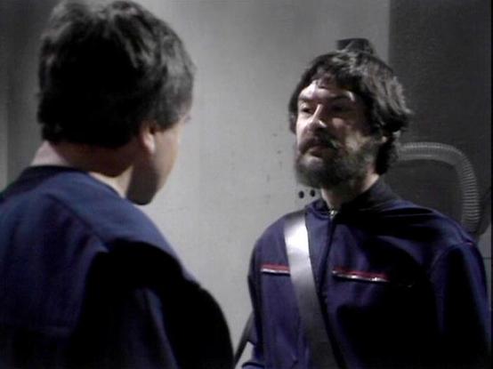 Claude Sonnet 4 describes the image as: "This image appears to be from a science fiction television production, likely from the 1970s or 1980s based on the visual style and production quality. The scene shows two figures in matching blue uniforms with distinctive red trim, suggesting they're part of the same organization or crew. The setting appears to be inside a spacecraft or futuristic facility, with sleek, minimalist design elements typical of sci-fi productions of that era.

The uniforms a…