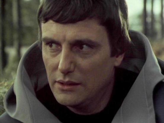 Claude Sonnet 4.5 describes the image as: "This image shows a scene filmed in a wooded outdoor setting with bare trees visible in the soft-focused background. The cinematography has a slightly muted, cool color palette typical of British television productions from the 1970s-80s era.

Paul Darrow appears in character wearing a distinctive costume featuring a light gray or silver padded vest or armor piece with prominent shoulder sections over a black underlayer. The costume has a science fictio…