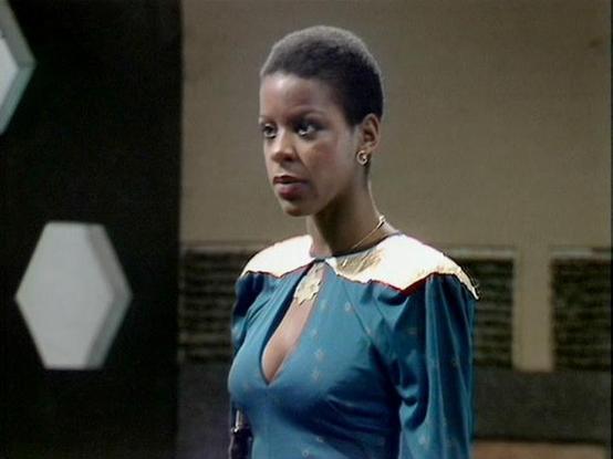 Claude Sonnet 4 describes the image as: "I can see this is a scene from what appears to be a science fiction television production, likely from the 1980s based on the aesthetic and production values. The setting appears to be a futuristic or space-based environment, with characteristic sci-fi set design visible in the background including geometric patterns and sleek surfaces.

The person in the image is wearing a distinctive blue garment with white collar details and decorative trim, which app…