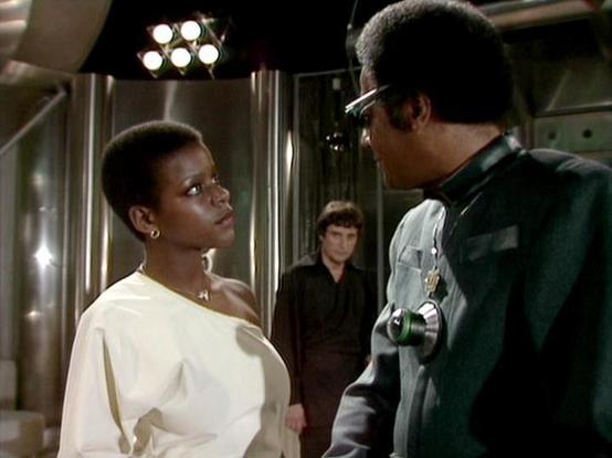 Claude Sonnet 4.5 describes the image as: "This image captures a tense dramatic moment from the British science fiction television series "Blake's 7." The scene takes place in what appears to be a spaceship interior, characterized by sleek metallic walls and futuristic lighting fixtures visible at the top of the frame.

In the foreground, two characters face each other in what seems to be a confrontational exchange. On the left stands a woman wearing an elegant white off-shoulder garment with g…