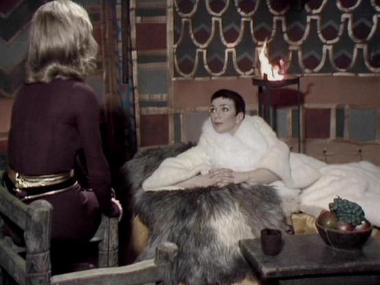 Claude Haiku 4.5 describes the image as: "# Scene Description

This appears to be a scene from a science fiction production, likely from the 1970s based on the visual style and color grading. The setting is an ornate interior space with decorative patterned panels visible in the background and what appears to be a fireplace or light source. The room features rich furnishings including cushioned seating and elegant drapery.

Two women are shown in conversation, with one seated on a bed or couch …