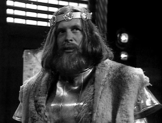Claude Sonnet 3.7 describes the image as: "This black and white image shows a medieval king or nobleman character from what appears to be a historical drama or film. The figure is wearing an ornate metal circlet or crown with decorative medallions across the forehead, and has long hair and a full beard. They're dressed in what looks like medieval royal attire including armor or a metallic breastplate with a fur-trimmed cloak or mantle draped over the shoulders.
The setting appears to be a dark…