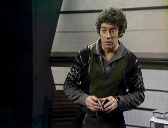 Claude Haiku 4.5 describes the image as: "# Image Description

This image shows a scene set in what appears to be a spaceship interior, characterized by the distinctive futuristic set design typical of 1970s science fiction television. The background features sleek, metallic paneling with horizontal lines and a modern architectural aesthetic.

The actor depicted is wearing a layered outfit consisting of a dark olive or brown tunic-style garment with a black leather jacket over it. The jacket fe…