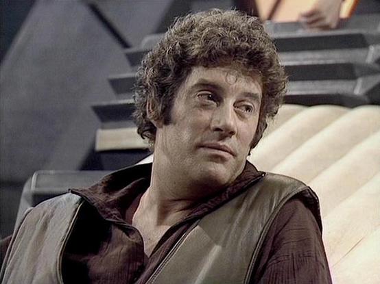 Claude Sonnet 4.5 describes the image as: "This image shows a scene from the British science fiction television series "Blake's 7," which aired from 1978 to 1981. The actor Gareth Thomas appears in character as Roj Blake, the series' protagonist and leader of a group of rebels fighting against a totalitarian interstellar federation.

In this shot, Thomas is seated in what appears to be the flight deck or control room of the Liberator, the advanced alien spacecraft that serves as the rebels' bas…