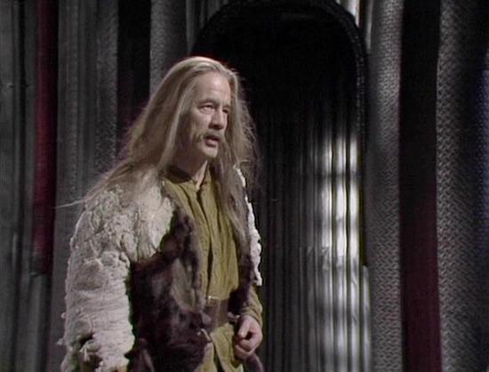 Claude Sonnet 4.5 describes the image as: "This image shows a dramatic scene from what appears to be a period television production. The actor is portrayed in elaborate historical costume, featuring long flowing blonde hair and layered garments including a yellowish scarf or neckerchief and a textured, weathered coat or cloak with fur trim. The clothing suggests a rustic or medieval setting, possibly depicting a vagrant, hermit, or someone from a lower social class in a historical drama.

The s…