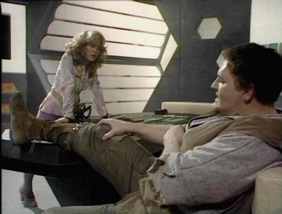 Claude Haiku 4.5 describes the image as: "# Scene Description

This image appears to be from a science fiction television production, set in a futuristic spacecraft interior. The scene depicts two characters in what looks like a command or recreational area of the vessel.

The setting features distinctive retro-futuristic design elements typical of 1970s sci-fi television, including geometric wall panels with hexagonal shapes and horizontal striping. The color palette consists of muted tones—gr…