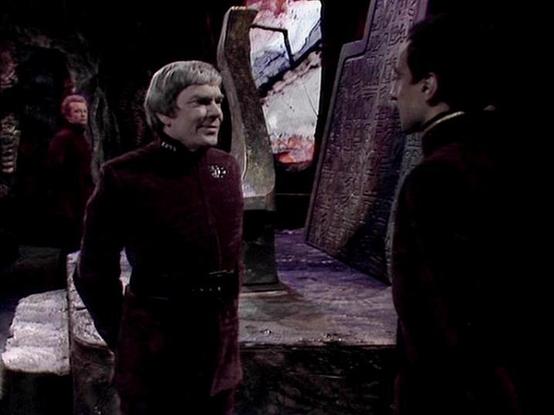 Claude Haiku 4.5 describes the image as: "# Scene Description

This appears to be a scene from Star Trek: The Next Generation, set in what looks like a Romulan vessel or similar sci-fi environment. The setting features dark, industrial architecture with large carved or textured panels in the background, creating an imposing alien atmosphere with purple and red lighting tones.

The man on the left wears a burgundy Starfleet uniform, characteristic of Starfleet officers from the TNG era. He appea…