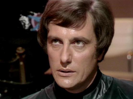 Claude Sonnet 3.7 describes the image as: "The image shows a close-up shot of a person with dark, short-styled hair and intense eyes looking slightly upward. They are wearing what appears to be a dark, possibly leather-like collar or jacket. The scene has a dramatic lighting quality typical of television productions from an earlier era, with a somewhat dark background that suggests an interior setting.

The image appears to be from a television production, likely from the 1970s or 1980s based o…