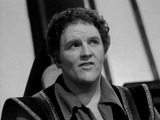 Claude Haiku 4.5 describes the image as: "I can see this is a black and white photograph of a man in what appears to be a television or film setting. He's wearing a distinctive embroidered or patterned jacket over a dark shirt, suggesting this may be from a science fiction production given the stylized costume design. The lighting and image quality suggest this is from a mid-to-late 20th century production.
The man has dark, curly hair and appears to be in the middle of speaking or reacting to…