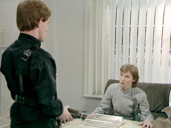 Claude Sonnet 4.5 describes the image as: "This image appears to be from a television production, showing a scene in an office setting with vertical blinds on the windows. The scene depicts two actors: a young man in a dark military or security-style uniform with buckle details standing with his back partially to the camera, and a woman seated at a desk wearing a light gray high-necked blouse or dress. The woman has short reddish-brown hair and is looking up at the standing figure with an atten…