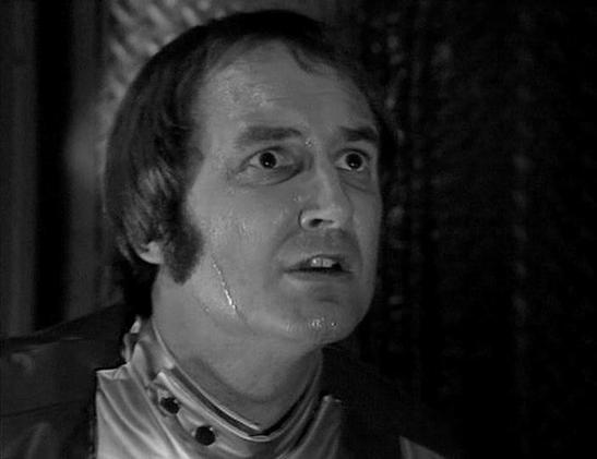 Claude Sonnet 3.7 describes the image as: "This is a black and white image from what appears to be a vintage television production. The image shows a person with distinctive sideburns wearing what looks like a high-collared costume or uniform with buttons visible at the neck. The individual has a surprised or concerned expression, with widened eyes and a slightly open mouth.

The setting appears dark and atmospheric with some textured background elements visible. The moody lighting and costume …