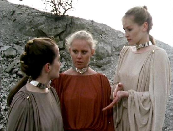 Claude Sonnet 4.5 describes the image as: "This image shows three actresses in a dramatic scene set against a barren, rocky landscape. The setting appears post-apocalyptic or otherworldly, with grey stone formations and sparse vegetation in the background.

The three women wear flowing, toga-like garments in earth tones - two in beige/grey and one in rust orange. They all sport distinctive metallic collar necklaces that suggest a futuristic or science fiction context. Their hair is styled simpl…