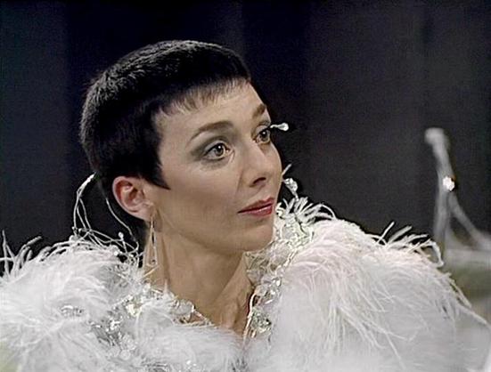 Claude Haiku 4.5 describes the image as: "# Image Description

This image shows a woman in what appears to be a scene from a classic science fiction television production, likely from the 1970s-1980s based on the styling and video quality. She wears an elaborate costume consisting of a white, feathered or textured garment with ornate embellishments and distinctive shoulder pieces adorned with decorative elements.

Her makeup is theatrical and dramatic, featuring bold eye makeup and defined feat…