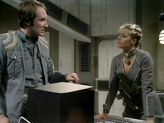 Claude Sonnet 4 describes the image as: "This image shows a scene from what appears to be a science fiction television production, set in a futuristic interior space with white/gray walls, ventilation grates, and industrial-style lighting. The setting has a distinctly spaceship or space station aesthetic typical of sci-fi shows from the late 1970s or early 1980s.

Two actors are positioned in the scene - a man on the left wearing a light-colored military or utility-style jacket, and a woman on …