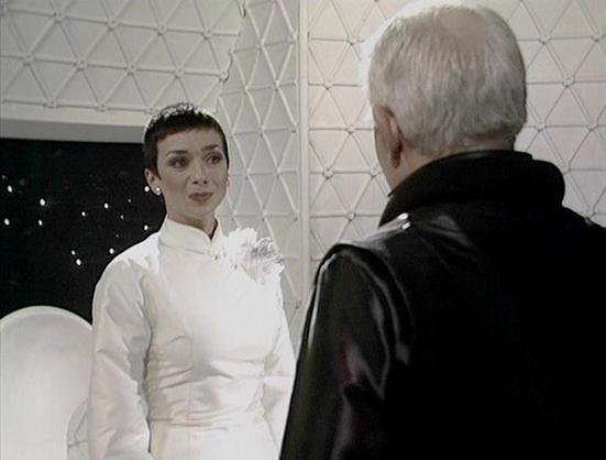 Claude Sonnet 3.7 describes the image as: "This image appears to be from a science fiction television production, likely from the British series "Blake's 7." The scene is set in what looks like a futuristic or spaceship interior with a distinctive geometric pattern on the white wall behind. 

The image shows two people in conversation. On the left is a person wearing an elegant white high-collared outfit with some decorative elements at the shoulder. Facing them is an older individual with whit…