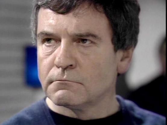 Claude Sonnet 4 describes the image as: "I can see this is a close-up shot of a middle-aged man with dark hair wearing what appears to be a dark blue or navy colored uniform or shirt. The image has the visual quality typical of British television from the 1970s or early 1980s, with that characteristic film grain and color saturation of the era. The lighting and framing suggest this is from a dramatic scene, possibly a science fiction or action television series. The man has a serious, contempla…