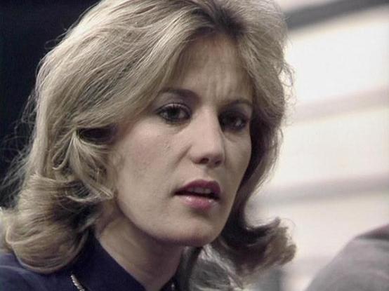 Claude Haiku 4.5 describes the image as: "# Image Description

This image shows a woman with shoulder-length blonde hair styled in a 1970s fashion, featuring bangs and waves. She has a serious, contemplative expression on her face, with her gaze directed downward. She's wearing what appears to be professional attire with a dark colored top or jacket and a delicate chain necklace.

The setting appears to be an indoor environment with neutral-colored walls in the background, suggesting an institu…