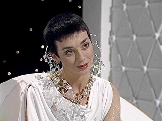 Claude Sonnet 3.7 describes the image as: "The image shows a person with short dark hair wearing an elegant white draped outfit or gown. They are adorned with an elaborate, statement necklace that appears to be made of clear or crystalline material. The setting looks like a stylized studio or film set with a textured geometric white background and some sparkling elements or lights visible. 

The costume has a classical or futuristic quality to it, with flowing fabric that drapes from the should…