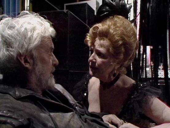 Claude Sonnet 4.5 describes the image as: "This appears to be a scene from a dramatic production, likely a film or television show. The image captures an intimate moment between two characters in what seems to be a dimly lit, indoor setting with dark walls and minimal lighting that creates a moody, theatrical atmosphere.
The scene shows two actors in close proximity, engaged in what appears to be an intense emotional exchange. The person on the left has distinctive white/grey hair and facial h…