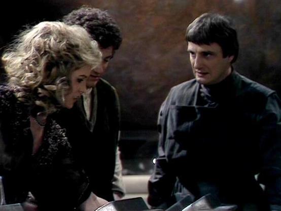 Claude Sonnet 4.5 describes the image as: "This image appears to be from the British science fiction television series "Blake's 7" (1978-1981). The scene shows three characters in what looks like a dimly lit spacecraft interior or similar enclosed setting. The composition features a blonde woman on the left wearing dark clothing with lace detailing, engaged in conversation with two men. One man stands in the middle background, while another is positioned on the right wearing a dark high-collare…