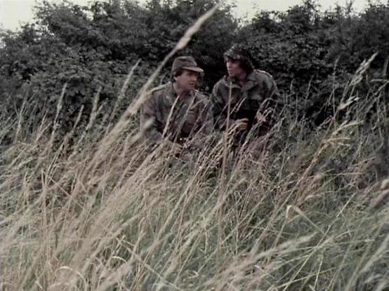 Claude Sonnet 4 describes the image as: "This image shows two people in military-style camouflage clothing positioned among tall grass in what appears to be a rural, countryside setting. The scene has the appearance of a military or wartime scenario, with dense hedgerows and vegetation visible in the background. The individuals are crouched or positioned low among the golden-brown grass, suggesting they may be in a reconnaissance or tactical situation. The lighting and overall aesthetic of the …