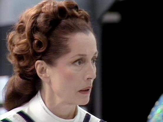 Claude Sonnet 4.5 describes the image as: "This image shows a woman in profile with an elaborate 1970s-style hairstyle featuring rolled curls on top and a long ponytail at the back. She has dark reddish-brown hair and is wearing what appears to be a white and dark-colored uniform or costume with a structured collar. The styling and aesthetic suggest this is from a science fiction television production, likely from the 1970s era based on the hair and makeup choices typical of that period's futur…