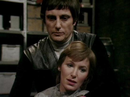 Claude Sonnet 4 describes the image as: "This appears to be a scene from a British science fiction television production, likely from the late 1970s or early 1980s based on the visual style and production values. The setting appears to be inside a spacecraft or futuristic facility, with characteristic metallic surfaces and storage compartments visible in the background.
The scene shows two characters in what appears to be an intimate or protective moment. One character wears a dark leather out…