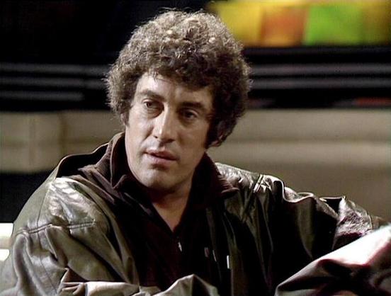 Claude Sonnet 4.5 describes the image as: "This image shows a scene from what appears to be a British science fiction television production, likely from the late 1970s or early 1980s. The actor wears a distinctive dark brown leather jacket over a black shirt, presenting a commanding presence. His curly hair and intense facial expression suggest he's in the middle of a dramatic moment or conversation.

The setting appears to be a futuristic interior, with soft lighting and what looks like colore…