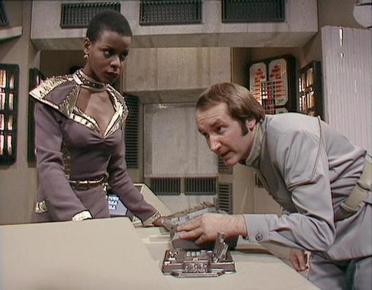 Claude Sonnet 4 describes the image as: "This image shows two actors in what appears to be a science fiction television production, likely from the late 1970s or 1980s based on the visual style and set design. The scene takes place in a futuristic control room or spacecraft interior, featuring typical sci-fi elements like control panels with blinking lights and a sterile, technological aesthetic.

One actor wears an elaborate costume with ornate metallic detailing and a high collar design that …