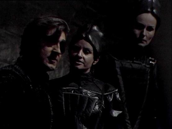 Claude Sonnet 3.7 describes the image as: "The image shows a dark scene from what appears to be a period film or television production. Three figures are visible in dim lighting, wearing what looks like medieval or Renaissance-style dark clothing with some leather elements and distinctive design features. The scene has a somber, dramatic atmosphere with the characters positioned close together in what might be a dungeon or castle interior.
The lighting is very low-key and atmospheric, creating…