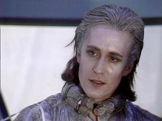 Claude Sonnet 4.5 describes the image as: "This image shows a scene from what appears to be a science fiction production, featuring distinctive theatrical makeup and costuming. The character has pale gray-toned skin and wears an elaborate period-style costume with ornate detailing around the collar area. The styling includes shoulder-length dark hair swept back from the face.
The makeup design creates an otherworldly appearance, suggesting an alien or non-human character. The costume includes …