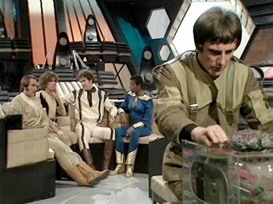 Claude Sonnet 4 describes the image as: "This appears to be a scene from the British science fiction television series "Blake's 7," set aboard the spacecraft Liberator. The image shows the ship's distinctive flight deck with its characteristic hexagonal wall panels and futuristic control surfaces. Several crew members are seated on the curved seating area in the background, wearing the show's typical beige/tan colored costumes. In the foreground, one crew member is operating what appears to be …