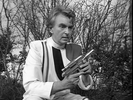 Claude Sonnet 4.5 describes the image as: "This black and white photograph captures a man in clerical attire outdoors in what appears to be a garden setting with bare branches visible in the background. He's wearing a white cassock with black trim and suspenders, styled as religious vestments. The subject holds what appears to be a wooden cross or crucifix in his hands, suggesting this is from a dramatic production with religious themes.

The composition places him against a natural backdrop of…