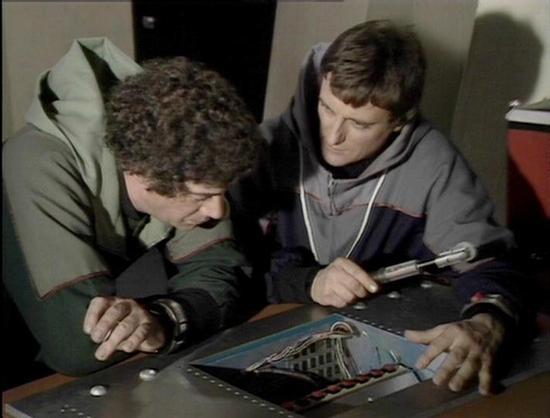 Claude Sonnet 4 describes the image as: "This appears to be a scene from the British science fiction television series "Blake's 7" from the late 1970s or early 1980s. The image shows two men in futuristic military-style clothing examining what appears to be a tactical display or map on a table. The setting looks like the interior of a spacecraft, with typical sci-fi production design elements visible in the background including control panels and metallic surfaces. The lighting and film quality…