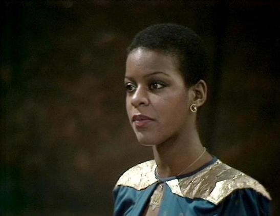 Claude Sonnet 3.7 describes the image as: "This image shows a person in what appears to be a period drama or theatrical production. They are wearing a distinctive costume with a blue dress or gown featuring a gold or metallic collar area. The person has short hair and gold earrings, and is photographed against a dark background that creates a dramatic effect.

The styling and costume suggest this is likely from a professional production, possibly from television or film from several decades ago…