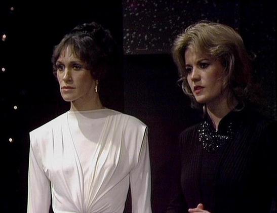 Claude Haiku 4.5 describes the image as: "# Scene Description
This image captures two women in what appears to be a science fiction setting, likely from a television production. The scene is set against a dark background with subtle lighting effects, creating an atmospheric, futuristic ambiance.
The woman on the left wears a flowing white garment with draped sleeves, her dark hair styled in a 1970s-inspired cut. She gazes to the side with a composed expression.
The woman on the right is dres…