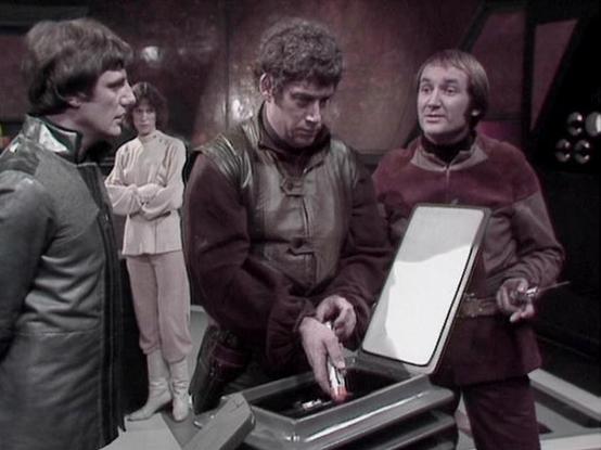 Claude Sonnet 4 describes the image as: "This appears to be a scene from the British science fiction television series "Blake's 7" from the late 1970s. The image shows several characters gathered around what appears to be a futuristic control panel or device with an open compartment. The setting has the distinctive low-budget but atmospheric look characteristic of BBC science fiction productions of that era, with curved walls and dim lighting creating a spaceship or underground base environment…
