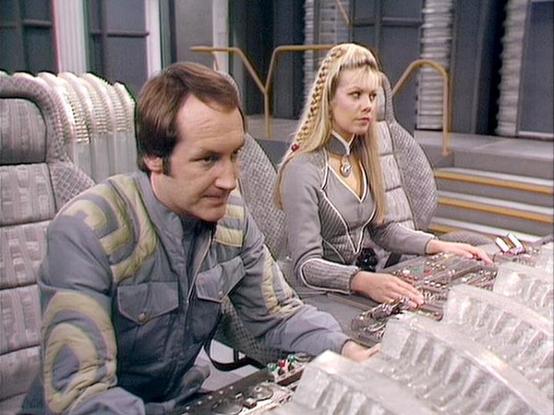Claude Sonnet 4.5 describes the image as: "This image shows a scene from what appears to be a science fiction television production, featuring two actors at a control panel in a futuristic spacecraft or facility setting. The male actor on the left wears a military-style olive and gray uniform with darker accents, while the female actor on the right has long blonde hair styled with braids and wears a form-fitting gray outfit with geometric quilted patterns and a distinctive necklace.

They are s…