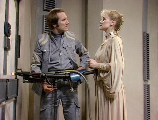 Claude Haiku 4.5 describes the image as: "# Scene Description

This image shows two figures in what appears to be a sci-fi setting, likely from a television production. The male character on the left wears a grey and gold uniform typical of 1970s space opera costumes, complete with practical pockets and fitted design. He's holding what appears to be a futuristic rifle with blue and black coloring.

The female character on the right wears a flowing, draped cream or beige garment with elegant ple…