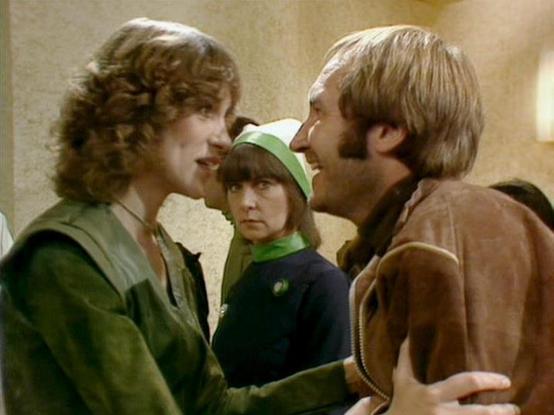 Claude Sonnet 3.7 describes the image as: "This image appears to be a scene from the classic British science fiction television series "Blake's 7" from the late 1970s/early 1980s. The scene shows three people in conversation, with the individual on the left wearing a green outfit with curly hair, another person in the middle wearing a green cap or hat looking somewhat disapproving, and a person on the right in a brown jacket with shorter hair. The setting appears to be an interior space with ne…