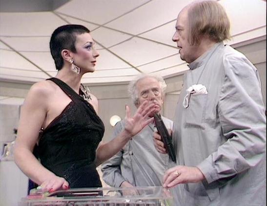 Claude Sonnet 4.5 describes the image as: "This image appears to be from a science fiction production, likely from the late 1970s or early 1980s based on the aesthetic and production design. The setting features a sleek, futuristic interior with geometric white panels and distinctive angular ceiling architecture characteristic of classic sci-fi television sets.

Three actors are engaged in what appears to be a tense dramatic scene. On the left, a woman with short dark hair wears an elegant blac…