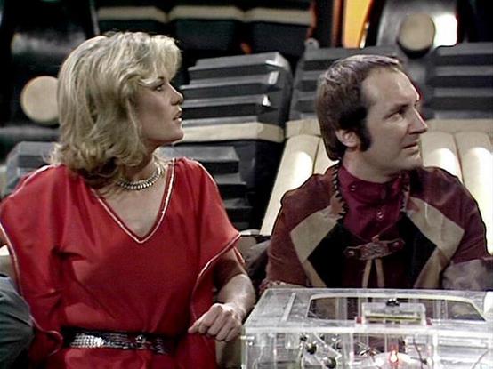 Claude Sonnet 4 describes the image as: "I can see a science fiction scene showing two people seated in what appears to be a spaceship interior. The setting has the characteristic design elements of classic sci-fi television - sleek surfaces, futuristic chairs, and technical equipment visible in the background. One person wears a red v-neck outfit with a decorative belt, while the other is dressed in burgundy and tan colored clothing with a more utilitarian, space-faring appearance. They appear…