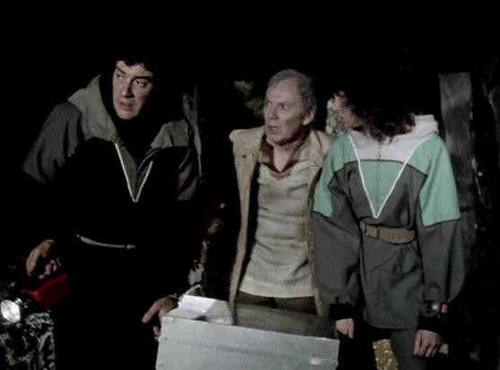 Claude Sonnet 4.5 describes the image as: "This image appears to be from a science fiction television production, likely from the 1970s based on the visual quality and costume design. The scene takes place in what looks like a dark, cave-like or underground setting with rough stone walls visible in the background.

Three figures are present in the frame, wearing distinctive futuristic uniforms characterized by two-tone color schemes with V-shaped chevron designs on the chest. The person on the …